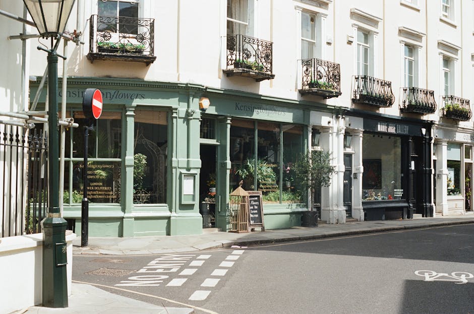 A street view in South Kensington showing a historic building with a ground-floor flower shop featuring large glass windows displaying potted plants and floral arrangements. The shop has a green facade with decorative columns and a black signboard for the flower shop, alongside a small chalkboard sign on the sidewalk listing specials. Above, the building has multiple white-framed windows adorned with black wrought iron balcony railings and flower boxes filled with greenery. To the left, there is a traffic sign mounted on a pole, and the street has painted markings indicating a bicycle lane and a pedestrian crossing. The scene is illuminated by natural daylight, with vehicles and pedestrians not visible, conveying a quiet, urban atmosphere typical of residential and commercial areas in central London, relevant to the context of house removals and furniture transportation as handled by companies such as South Kensington Movers.