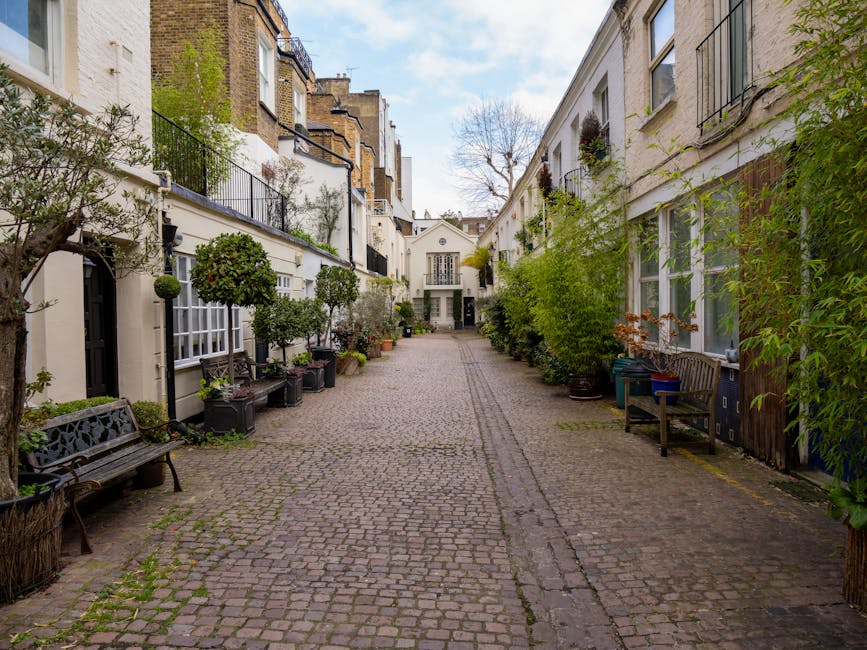 A cobblestone courtyard in a residential area within Kensington, featuring white-painted brick houses on both sides with black metal balconies and window frames. The courtyard is lined with potted pla