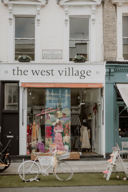 The image depicts the exterior of a shop on Fulham Road, a busy commercial street in South Kensington, during daytime with natural lighting. The storefront features large glass windows displaying colorful clothing, including a prominently positioned pink dress with floral patterns, along with other garments and decorative items arranged on mannequins and display stands inside. Above the entrance, there is a sign reading 'the west village' and an awning with the website address 'www.westvillage.com' in pink text. In front of the shop, a white bicycle is parked on the sidewalk, partially covered with a clear plastic wrap, suggesting recent or ongoing packing for a move or storefront rearrangement. To the right of the storefront, a teal-colored building with a small awning can be seen, and nearby, a white picnic table is positioned on a patch of grass. The environment appears to involve a commercial setting where furniture and merchandise are temporarily housed outside, possibly as part of a customer or business relocation process, with South Kensington Movers periodically handling furniture transport or packing and moving activities from or to this street-level shop. The brickwork of the upper floors and window details hint at the historic architectural style typical of the area.