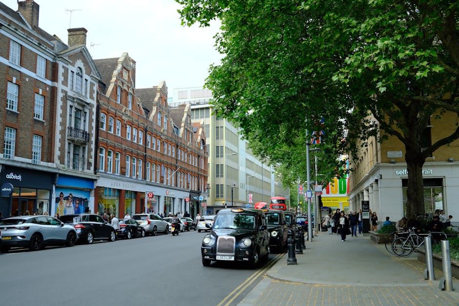 A busy street scene in South Kensington during daytime, showing a row of traditional brick and stone commercial buildings with retail stores on the ground floor, including a clothing shop and a self-portrait gallery. Several parked cars, including a London black cab, line the street, with some vehicles waiting or moving along the road. Pedestrians walk along the wide sidewalk, which is shaded by large, leafy green trees. Bicycles are secured to bike racks near the storefronts, and bollards separate the pavement from the street, facilitating loading and unloading activities. The lighting is natural with overcast skies, creating a neutral environment indicative of typical city street scene. This setting reflects elements involved in urban furniture transport, moving logistics, and packing processes, relevant to house and commercial relocation services like those provided by South Kensington Movers, as seen on their website page for Fulham Road commercial removals for shops and galleries.