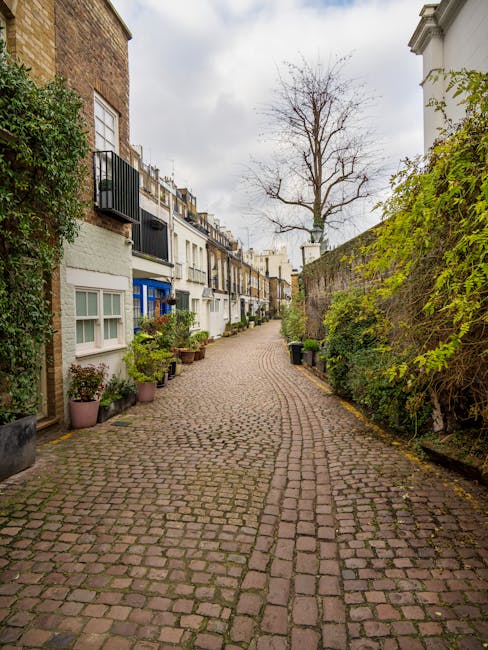 A narrow residential street paved with small cobblestones, lined with closely spaced terraced houses on the left side featuring white and blue facades, some with small front gardens decorated with potted plants and shrubs. The houses have varied window styles, with some bay windows and balcony railings. On the right side, a tall brick wall covered with green climbing plants separates the street from neighboring properties. In the center of the image, a leafless tree extends upward towards an overcast sky. The street appears quiet and is suitable for home relocation or moving services such as furniture transport or packing and loading detailed by South Kensington Movers. The scene emphasizes the confined urban environment typical of South Kensington's narrow streets, often requiring specialized logistics for house removals.