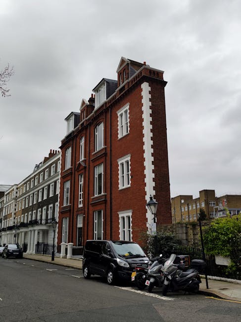 A multi-storey red brick building with white window frames and decorative white accents along the edges, situated on a residential street under an overcast sky. In front of the building, a black van and several scooters are parked on the pavement. The street features a row of similar Victorian-style terraced houses with white facades and black railings. Some of these buildings have bay windows and small front gardens enclosed by low walls. The image captures a typical urban setting potentially subject to house removals or furniture transport due to the presence of parked vehicles and the overall streetscape, aligning with professional moving services like those provided by South Kensington Movers for narrow street home relocations.