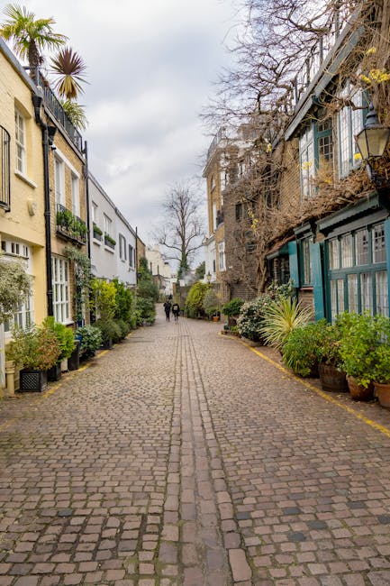 A narrow residential street paved with small cobblestones, lined with closely spaced terraced houses on the left side featuring white and blue facades, some with small front gardens decorated with potted plants and shrubs. The houses have varied window styles, with some bay windows and balcony railings. On the right side, a tall brick wall covered with green climbing plants separates the street from neighboring properties. In the center of the image, a leafless tree extends upward towards an overcast sky. The street appears quiet and is suitable for home relocation or moving services such as furniture transport or packing and loading detailed by South Kensington Movers. The scene emphasizes the confined urban environment typical of South Kensington's narrow streets, often requiring specialized logistics for house removals.