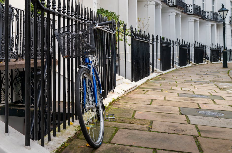 A blue bicycle with a black front basket is secured to a black metal fence outside a residential property in South Kensington. The bicycle is leaning against the fence, which runs along a paved sidewalk adjacent to a white building with architectural columns and a staircase leading to the entrance. Several black fencing panels extend along the pavement, with some white stone steps behind the fence. The pavement consists of large, irregularly shaped stone slabs with small gaps between them, some of which contain moss or grass. The scene is well-lit, indicating daytime, and the setting reflects a typical urban residential street suitable for house removals and furniture transport services. South Kensington Movers operate in this area, often assisting with packing, loading, and relocating household furniture within such environments, respecting council rules for removals and waste disposal.