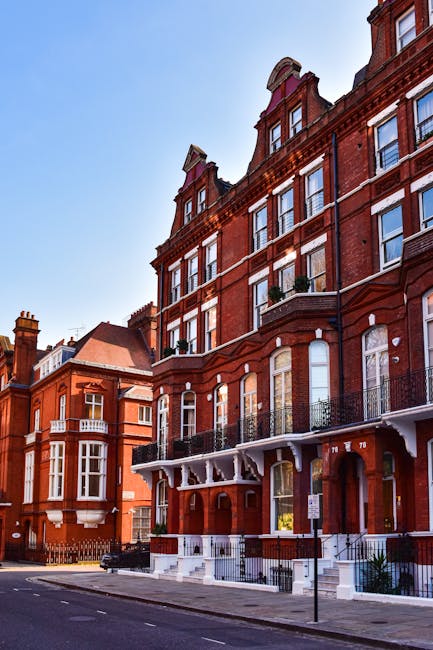 Three-storey red brick residential buildings with ornate white decorative features on the ground floor and along the roofline, situated on a paved city street. In the foreground, a black van is parked beside the pavement, with a loading area adjacent to the buildings where furniture and large cardboard boxes are being unloaded. Visible items include a wooden dining table with chairs, wrapped in plastic, and several cardboard boxes, some secured with packing tape and plastic wrapping, placed on dollies or being carried by movers. A trolley with a strap is used to help load the furniture into the van, indicating an active home relocation process. The buildings have small balconies with wrought iron railings on the second floor, and the window openings feature white frames contrasting with the brickwork. Bright daylight illuminates the scene, showcasing the movement of household items during the loading process, consistent with professional removals services such as those offered by South Kensington Movers, especially relevant to discussions on parking permits and logistics for house removals in Kensington.