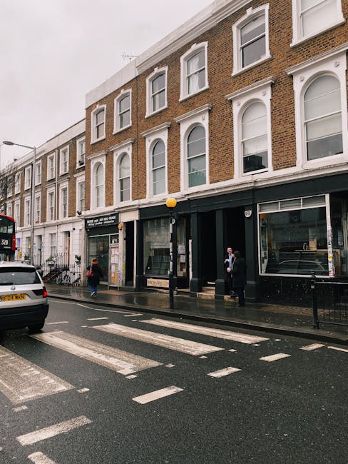 Street view of a multi-storey brick building with large display windows on the ground floor, located on a wet pavement during overcast weather. The building has white-framed arched windows on the upper floors and a black storefront entrance with glass doors. In front of the building, a black bollard with a yellow top is situated on the sidewalk, alongside a small group of people, including a person wearing a dark suit and mask, and another individual in casual clothing. A white van and a black car are parked on the street, which features a zebra crossing with white striped markings, and there are bicycles leaning against the building. This scene appears to be part of a home relocation or furniture transport process, supported by the presence of people possibly preparing for or concluding a move, with South Kensington Movers providing the professional removals services in the area, as related to the Kensington High Street movers tips for tight access properties.