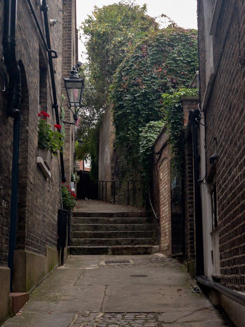 A narrow cobbled stairway leading uphill between two brick buildings in a residential area, with external black metal railings on one side and a small wall with greenery and climbing plants on the oth