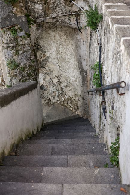 A narrow cobbled stairway leading uphill between two brick buildings in a residential area, with external black metal railings on one side and a small wall with greenery and climbing plants on the oth