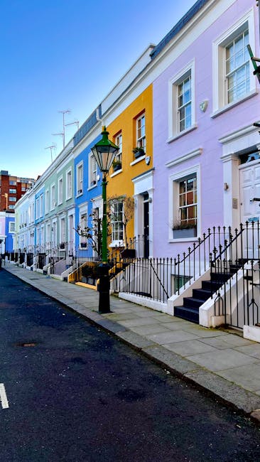A row of colorful terraced houses in Kensington with pastel facades in shades of blue, yellow, purple, and white, each featuring white-framed windows, black wrought-iron railings along small front steps, and traditional lampposts positioned on the pavement in front. The buildings are illuminated by natural daylight, highlighting the clean, well-maintained exteriors. To the side, a narrow street with a dark asphalt surface runs parallel to the houses. Inside one of the houses, visible through an open doorway, there are cardboard moving boxes, plastic wrapping, and packing materials indicating preparations for a home relocation. On the pavement near the entrance, a few moving blankets and hand trucks are positioned, with the ongoing loading process suggesting furniture transport as part of a professional removals service. The environment reflects a typical residential area in South Kensington, where South Kensington Movers offer removals and packing services for house moves, emphasizing the logistical aspects of furniture transport and home packing.