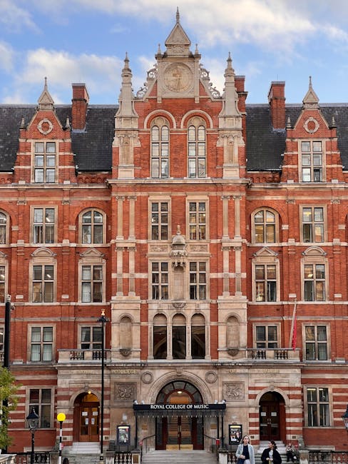 Photograph of the Royal College of Music building in South Kensington, featuring an ornate red brick façade with stone detailing, tall arched windows, and decorative spires on the rooftop. In front of the building, a moving team from South Kensington Movers is engaged in a home relocation process, loading cardboard boxes, wrapped furniture, and belongings onto a van parked adjacent to the pavement. Visible equipment includes hand trucks, straps, and moving blankets protecting the furniture during the transport. The scene captures the interior of the entrance area with a large doorway, set against a clear sky, illustrating the logistics involved in professional furniture transport and packing for a house move or cultural institution relocation, consistent with the themes of removals and moving services linked to Royal Albert Hall landmark moves advice.